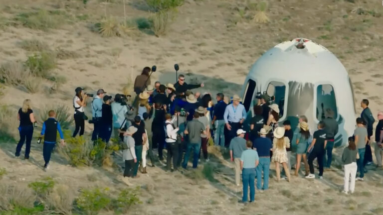 A group of people gathers around a landing capsule in a desert area. Some individuals appear to be assisting or inspecting the capsule, while others are taking photographs and observing.