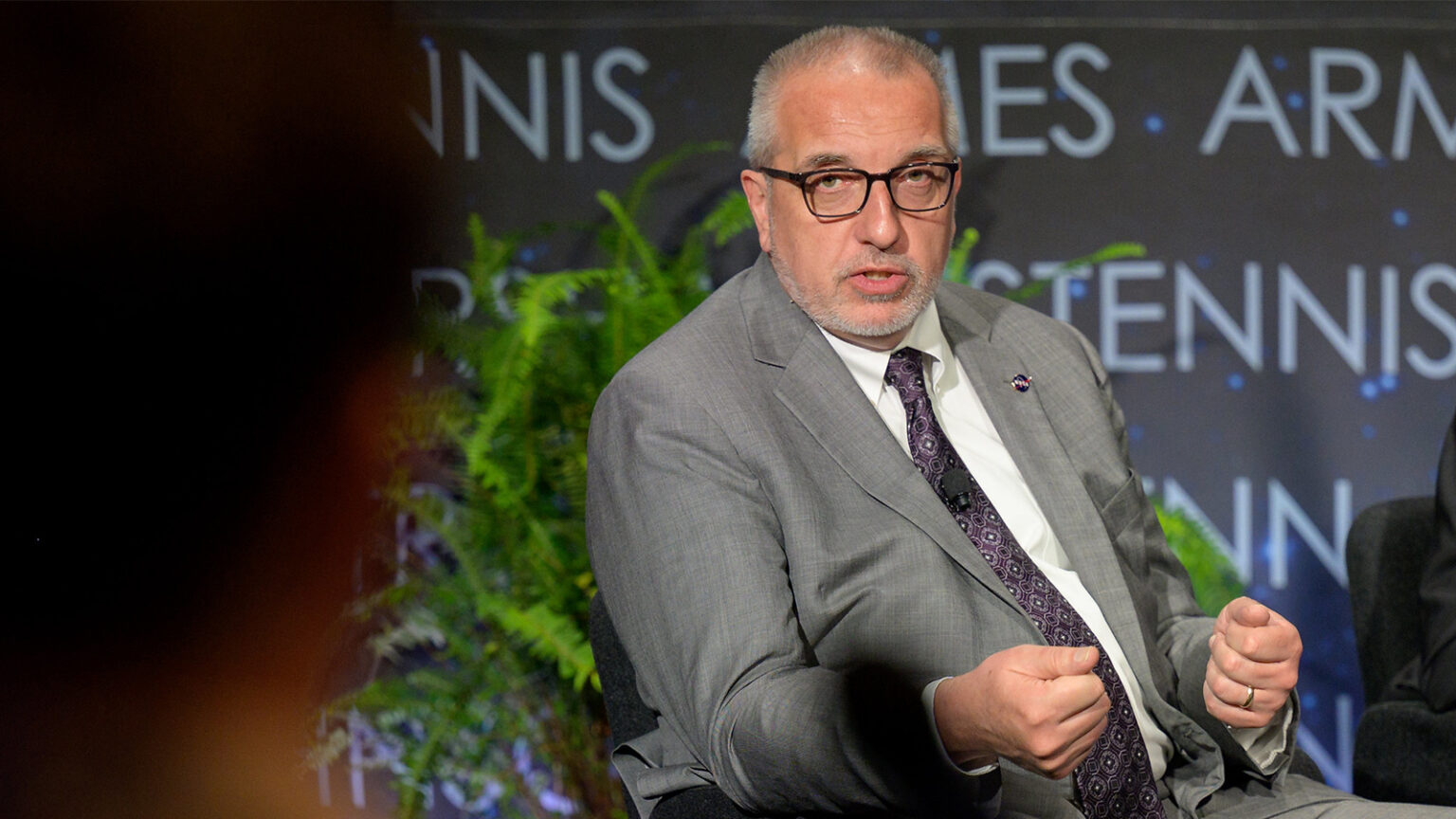 A man in a gray suit and glasses speaks while seated, with a potted plant and a partially visible person in the foreground.