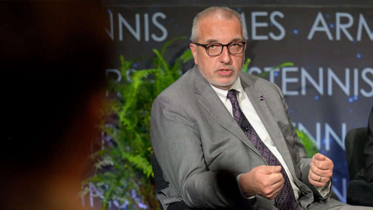 A man in a gray suit and glasses speaks while seated, with a potted plant and a partially visible person in the foreground.