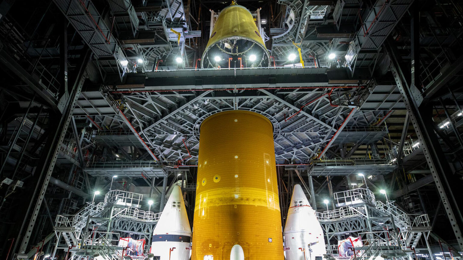 A large rocket component is centered indoors, surrounded by scaffolding and other space components, under bright lights inside an industrial facility.