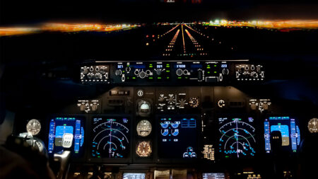 The image shows the illuminated cockpit of an aircraft during nighttime, with the runway lights visible through the windshield as the plane approaches for landing.
