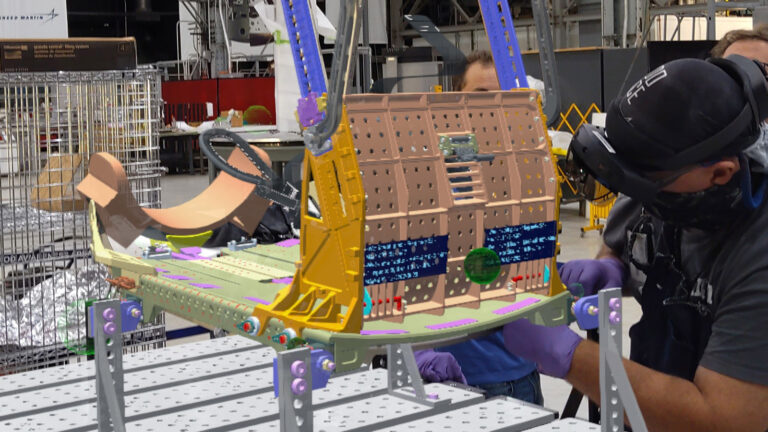 Technicians work on assembling a complex mechanical structure in an industrial setting, wearing protective gear and using specialized equipment.