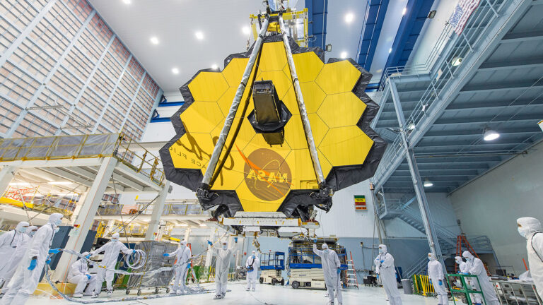 Scientists in cleanroom suits surround the James Webb Space Telescope, which features a large segmented gold mirror, during final preparations in a high-tech facility.
