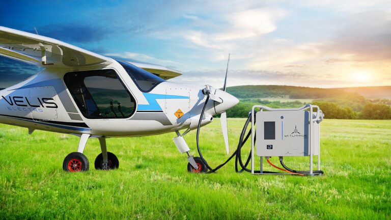 A small electric airplane is parked on a grassy field, connected to a charging station. The sky is partly cloudy with the sun low on the horizon.