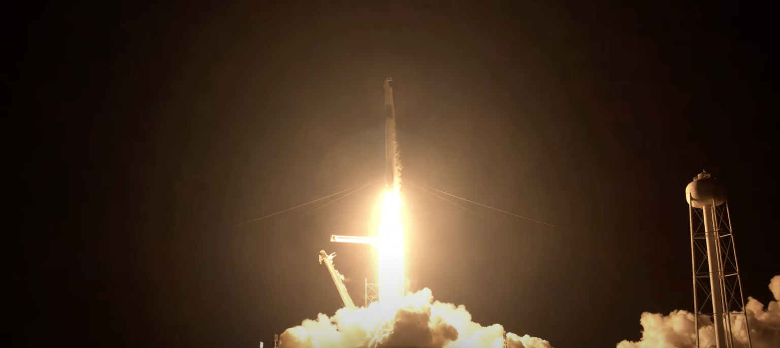 A rocket is launching at night with a bright flame and smoke cloud. The launch pad and support structures are visible.