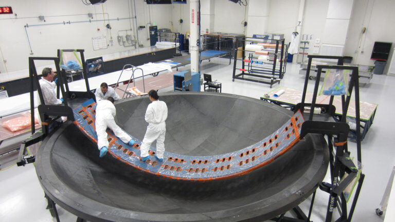 Three people in white protective suits work on a large, curved carbon fiber structure in an industrial cleanroom.
