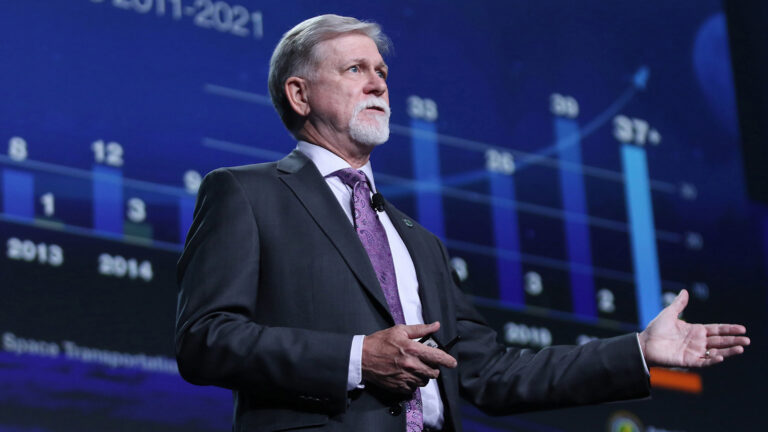 A man in a suit and tie is speaking on stage with a projected bar chart showing data from 2011 to 2021 in the background.