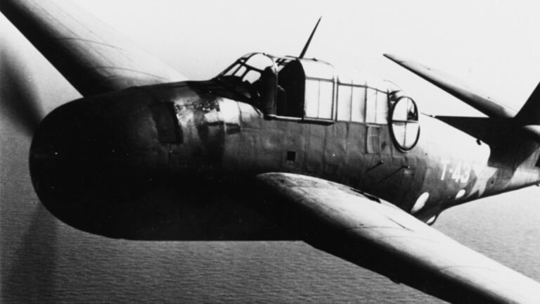 Close-up of a World War II-era military aircraft in flight, showing detailed markings and cockpit. The aircraft is flying over a body of water.