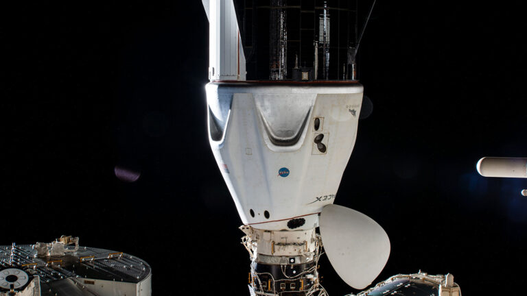 A spacecraft is docked at a space station module in the darkness of space. The craft has a white exterior with visible connection points and a NASA logo on it.
