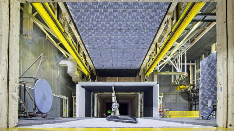 Interior view of a large industrial wind tunnel with intricate machinery, acoustic tiles, and a model aircraft being tested in the center.