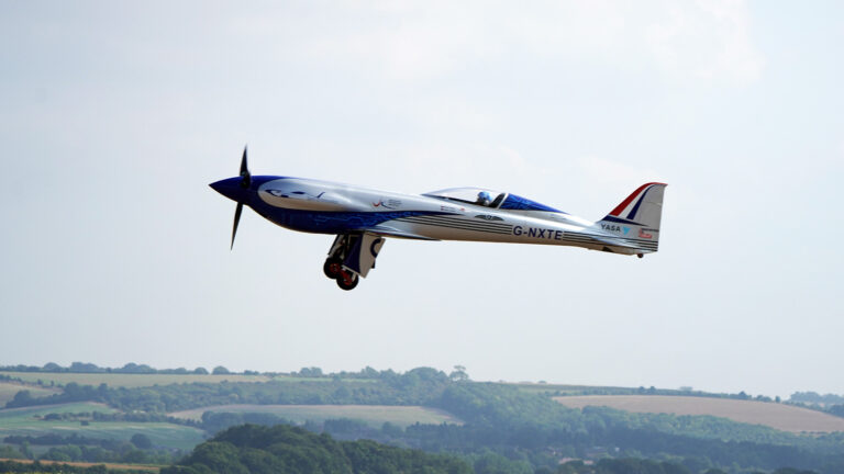 A single-engine propeller aircraft with registration number G-MXTE is flying over a landscape with rolling hills and sparse vegetation.