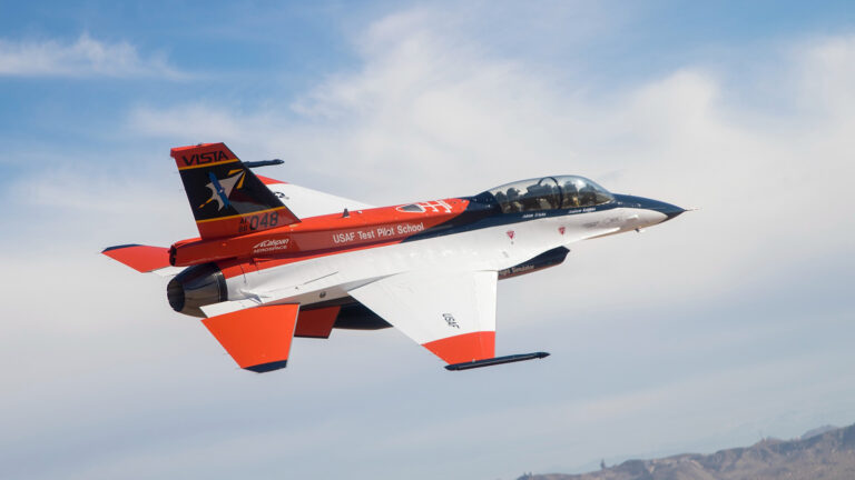 A single-engine jet aircraft with red, white, and black markings is in flight against a cloudy sky. The aircraft is labeled with "USAF Test Pilot School" and "VISTA.