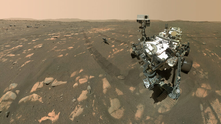 Mars rover and helicopter on the rocky surface of Mars under a dusty orange sky. The rover has visible tire tracks behind it.