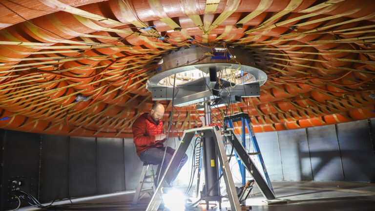 A person in a red jacket is seated on a stool inside a large circular structure, working on equipment attached to the ceiling using various tools and ladders.