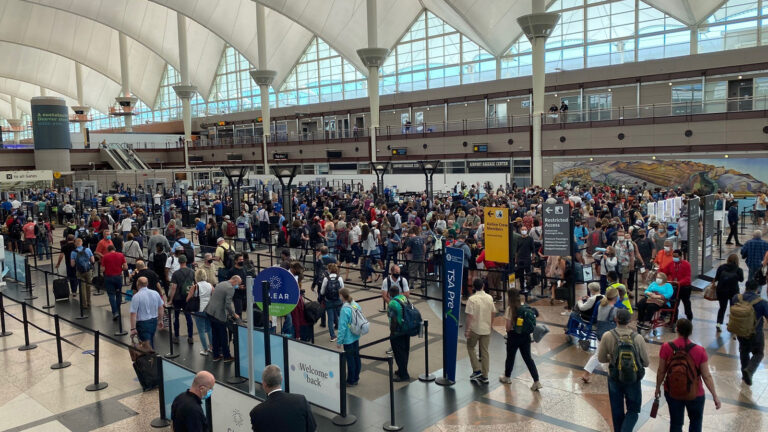 A crowded airport security checkpoint with numerous travelers in line, large windows in the background, and signage for different security lanes visible.