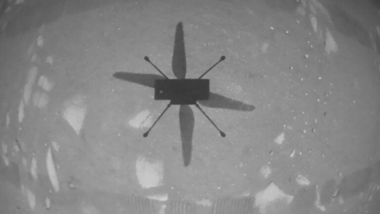 Black-and-white image of a quadcopter helicopter casting a shadow on a rocky, cratered surface below, likely on a planetary body or moon.