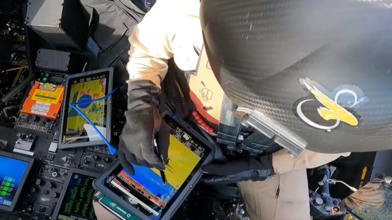 A person in a helmet and gloves operates a touchscreen device with a stylus, surrounded by various electronic equipment inside a cockpit.