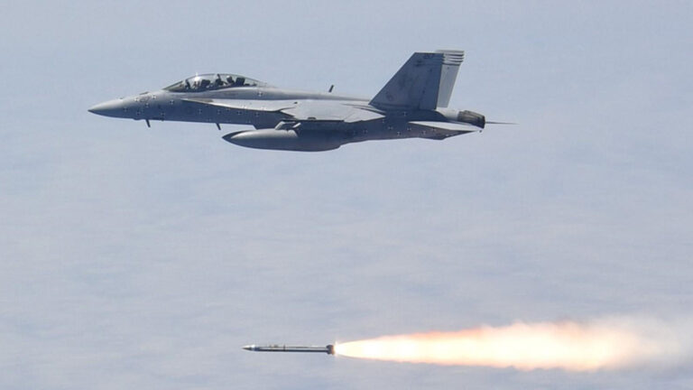 A fighter jet in flight releases a missile, which trails a bright flame as it speeds away from the aircraft.