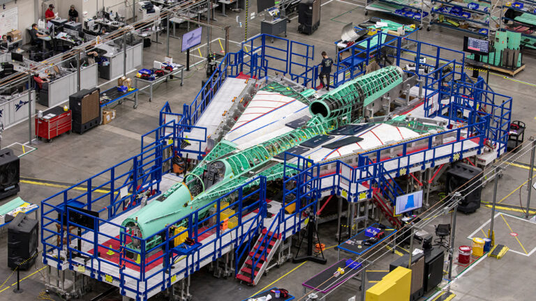 An overhead view of a partially constructed aircraft surrounded by scaffolding and equipment in an industrial facility with workers in the background.