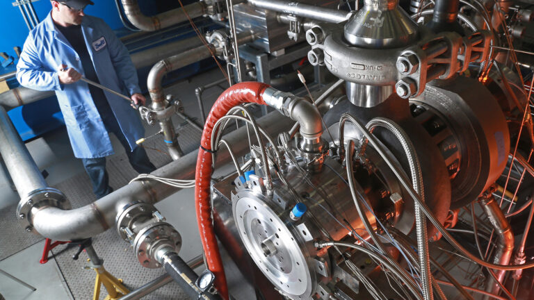 Progress in carbon-neutral technologies for power generation A person in a blue coat and cap is working on machinery with numerous pipes, wires, and metallic components in an industrial setting.