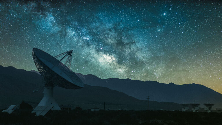 A large radio telescope dish is pointed toward a star-filled night sky, with a mountainous landscape in the background.