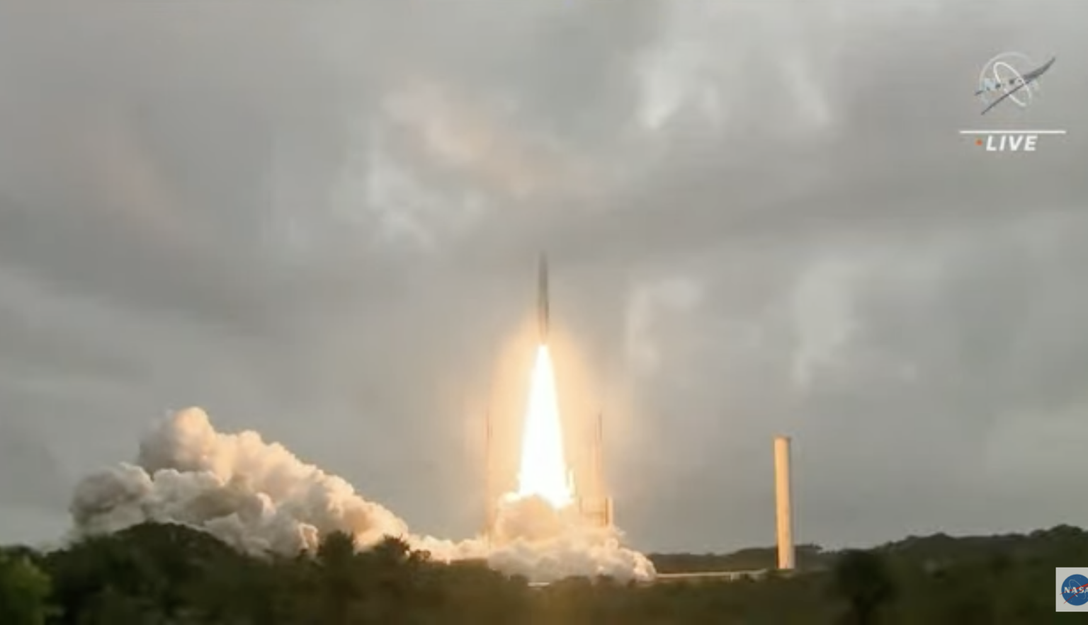 A rocket launches into a cloudy sky with flames and smoke trailing. A tall, thin structure stands nearby. The NASA logo is visible in the top right corner.