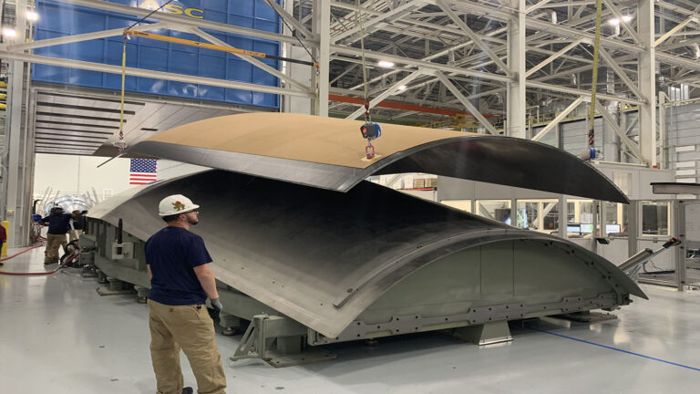 The right stuff for rocket joints A factory worker observes the assembly of large curved metal components in a spacious industrial facility. The U.S. flag is visible in the background, along with structural beams and machinery.