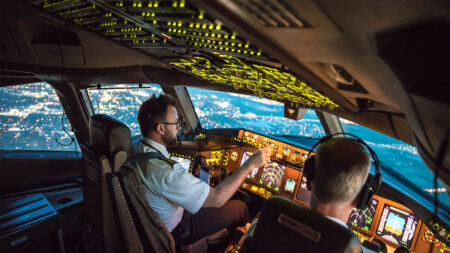 Two pilots, one in uniform, navigate a commercial airplane over a city at night, illuminated cockpit controls and exterior lights visible through the windows.