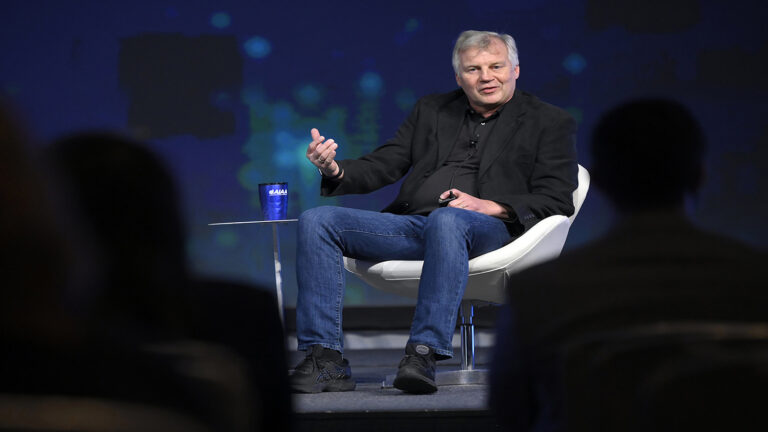 A man in a black blazer and jeans sits on a white chair on stage, speaking to an audience.