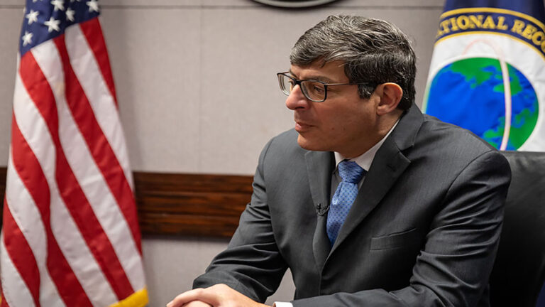 A man in a suit and tie sits in a room with an American flag and a seal on the wall in the background.