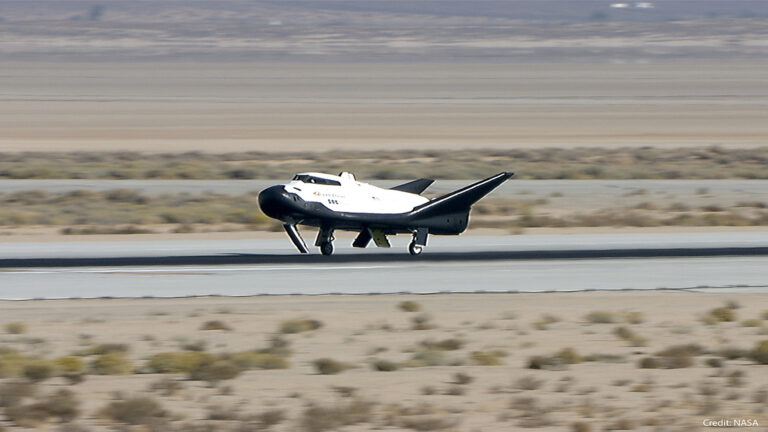A small futuristic-looking spacecraft with a white body and black wings is landing on a runway in a desert landscape. Credit: NASA.