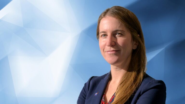 A woman with long brown hair is smiling slightly, wearing a dark blue blazer, standing in front of a geometric blue and white background.