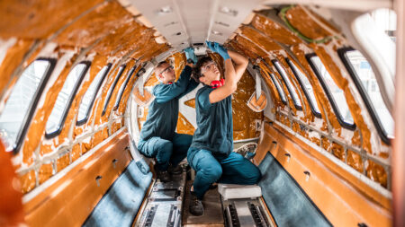 Two mechanics work inside the stripped-down fuselage of an aircraft, performing maintenance tasks while wearing green uniforms.