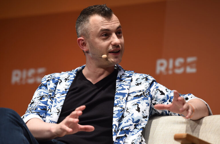 Person with a microphone headset speaking while seated on a stage with an orange background and "RISE" text.