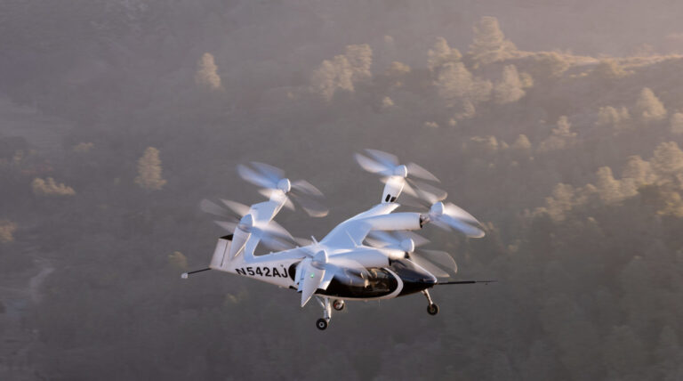 A white vertical take-off and landing aircraft with six rotors is flying over a forested mountainous area during sunrise or sunset.
