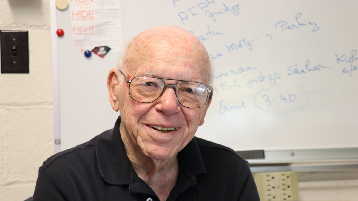 Elderly man with glasses smiling, seated indoors in front of a whiteboard with handwritten notes.