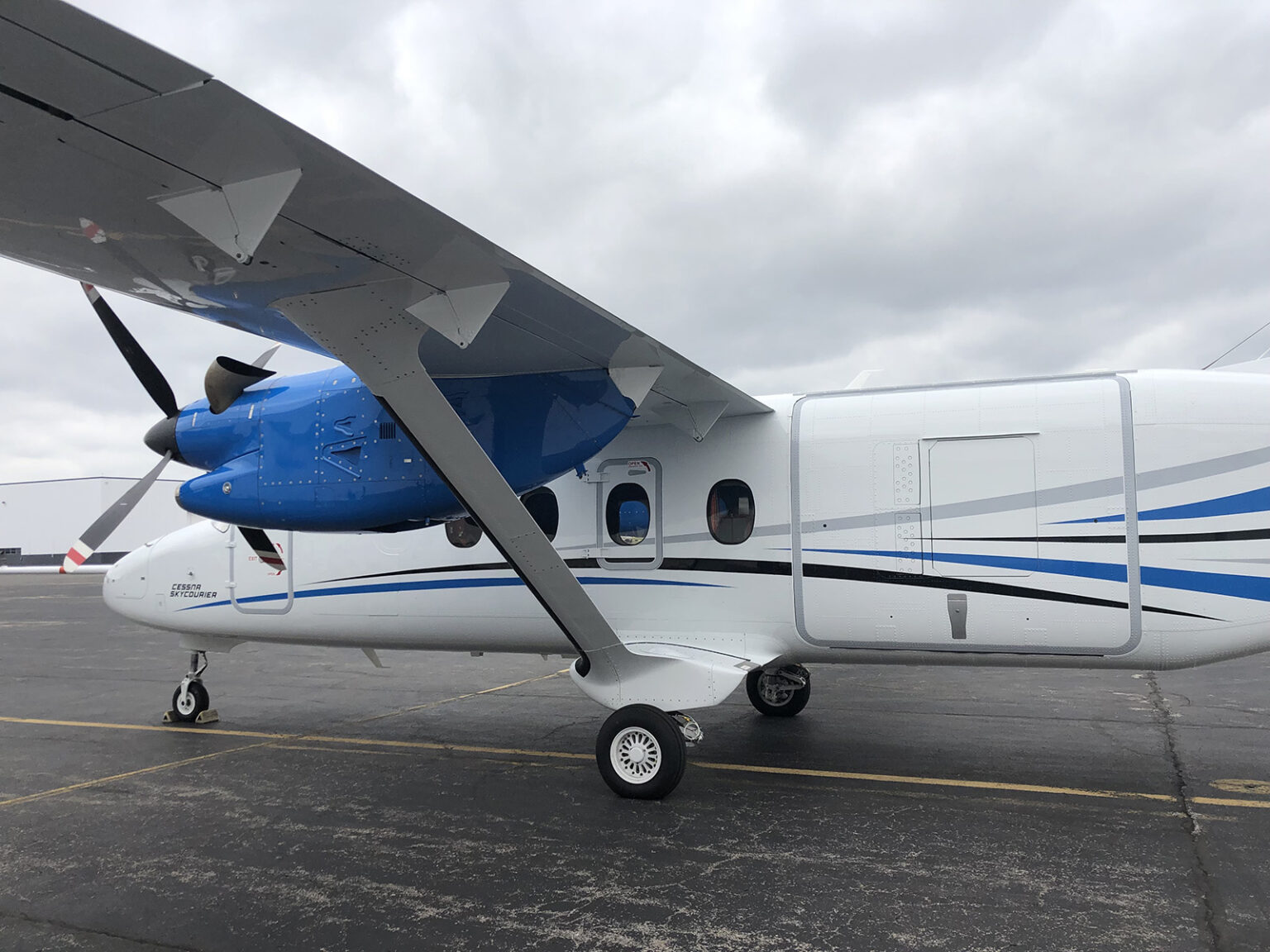 A small twin-engine aircraft parked on an airport tarmac under a cloudy sky. The plane is white with blue accents and has its propellers at rest.