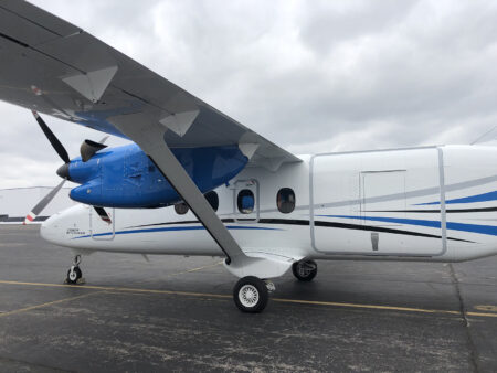 A small twin-engine aircraft parked on an airport tarmac under a cloudy sky. The plane is white with blue accents and has its propellers at rest.