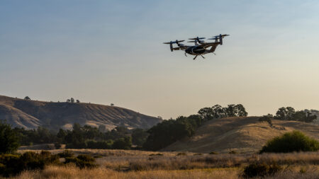 A drone with six rotors flies above a grassy, hilly landscape under a clear sky.