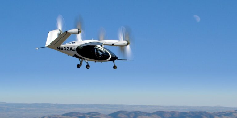 A white and black helicopter-like aircraft with the registration number N542AJ flies in a clear blue sky, with a mountainous landscape and a crescent moon visible in the background.