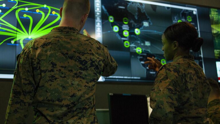 Two military personnel in camouflage uniforms stand in front of large screens displaying maps and data, discussing information or strategy.