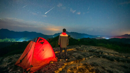 Person standing next to a lit tent on a mountain at night, looking at a sky filled with stars and shooting meteors.