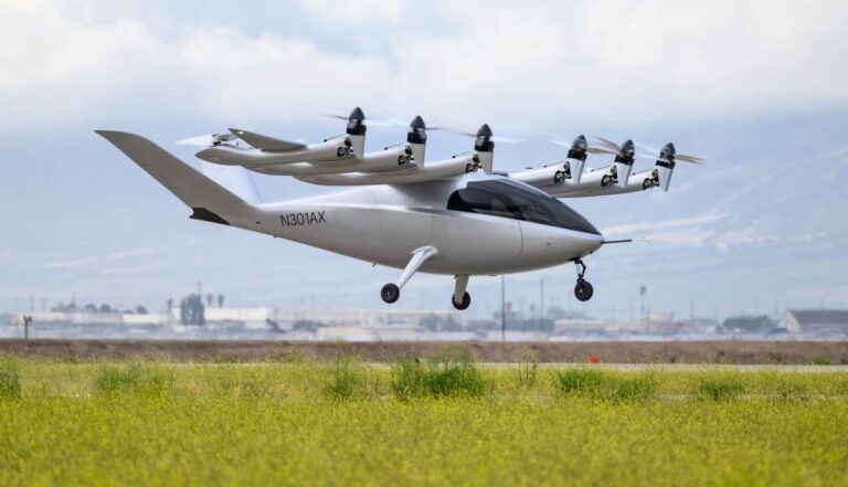 A white electric vertical takeoff and landing (eVTOL) aircraft with multiple propellers hovers above a grassy field.