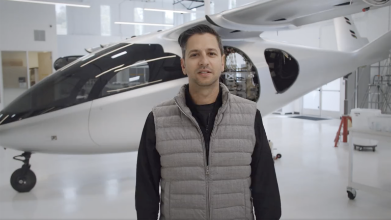 A man in a gray vest stands in front of a modern white aircraft in a spacious hangar.