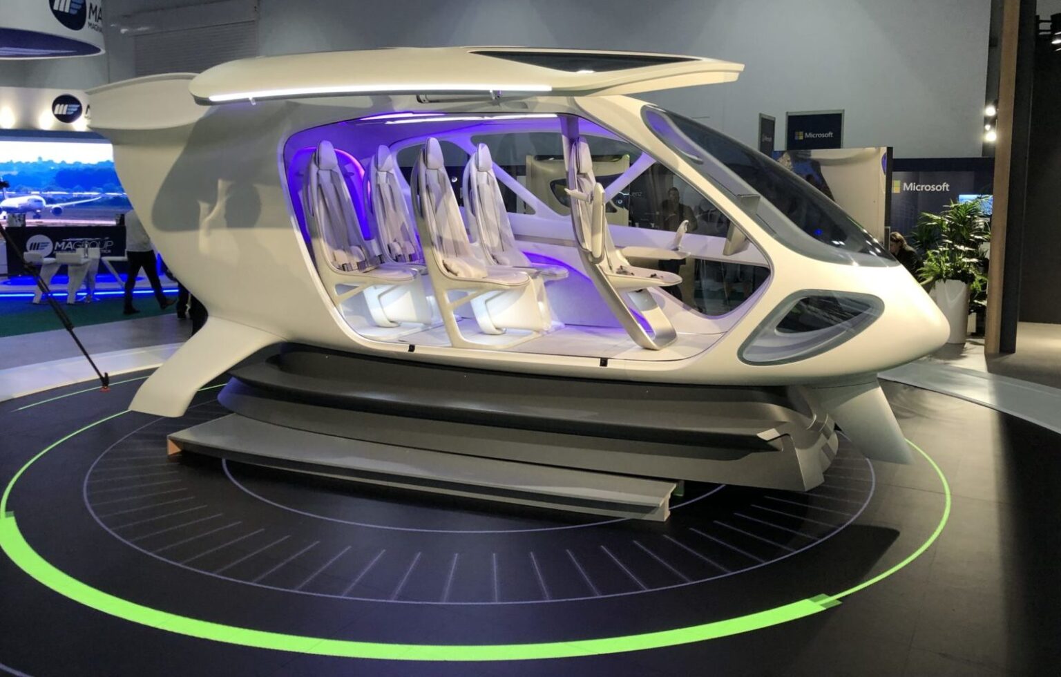 A futuristic, white, multi-seat flying vehicle prototype is displayed in an exhibition hall, illuminated with purple lighting inside and shown on a circular platform with green markings.