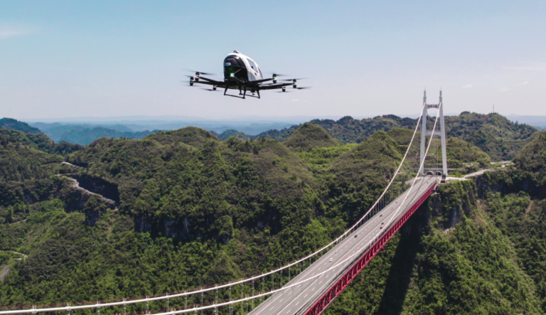A drone flies over a large cable-stayed bridge spanning a lush, green mountainous area on a clear day.