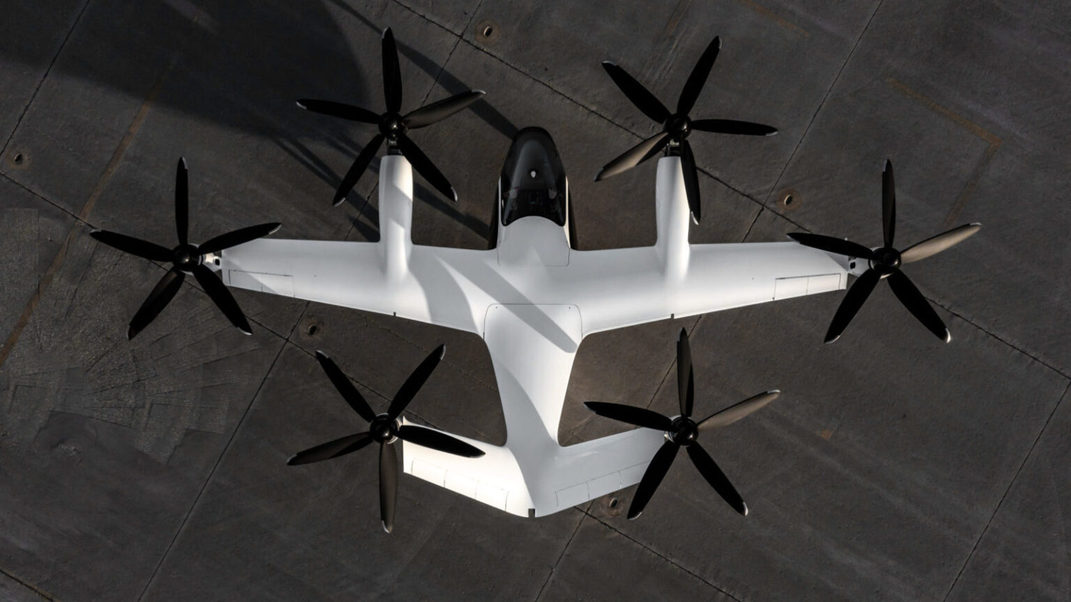 Aerial view of a white, six-rotor drone aircraft on a concrete ground. The drone has two large wings and a sleek, aerodynamic body.