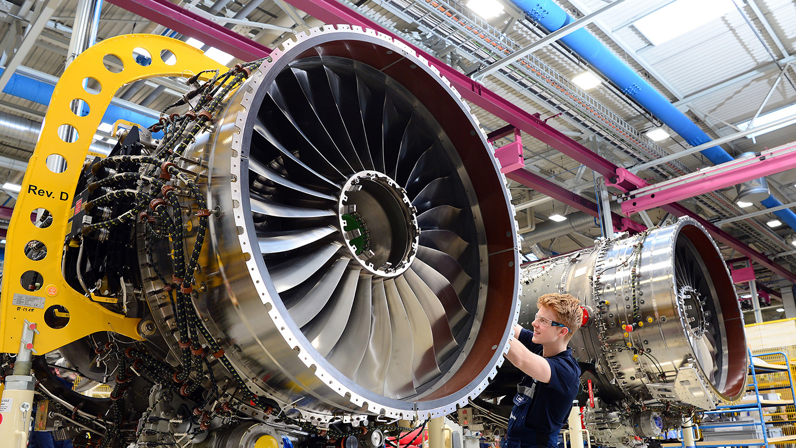 An engineer works on the large turbine of an aircraft jet engine in an industrial facility. Another similar engine is visible in the background.