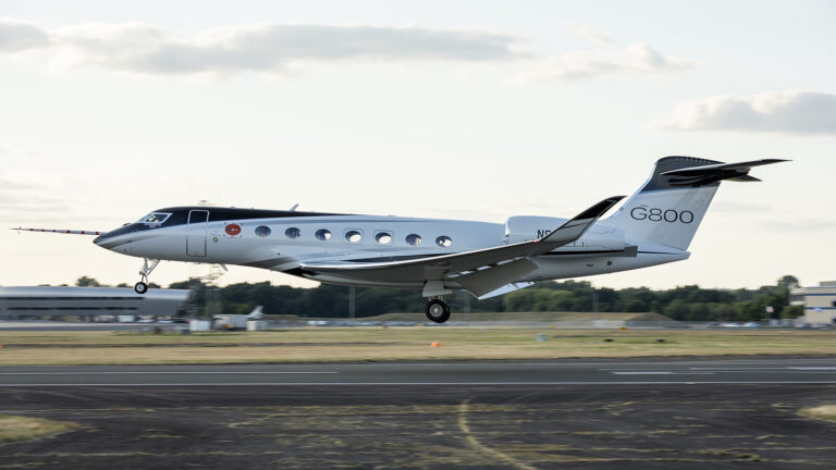 A Gulfstream G800 private jet lands on a runway during daylight, with buildings and trees visible in the background.