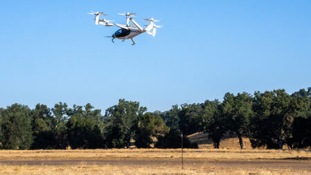 A white and black drone with six propellers flies over a dry, grassy field with trees in the background under a clear blue sky.